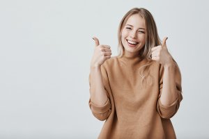 Portrait of fair-haired beautiful female student or customer with broad smile, looking at the camera with happy expression, showing thumbs-up with both hands, achieving study goals. Body language