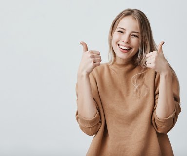 Portrait of fair-haired beautiful female student or customer with broad smile, looking at the camera with happy expression, showing thumbs-up with both hands, achieving study goals. Body language