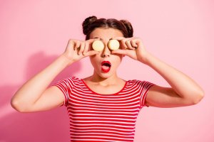 Close-up portrait of nice cute charming attractive glamorous lovely funny comic teen girl in striped t-shirt covering eyes with colorful delicious snack opened mouth isolated over pink background
