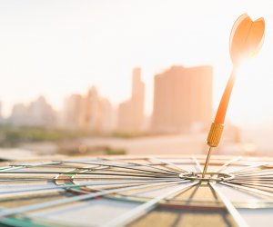 Red dart arrow hitting in the target center of dartboard with modern city and sunset background. Target business, achieve and victory concept .